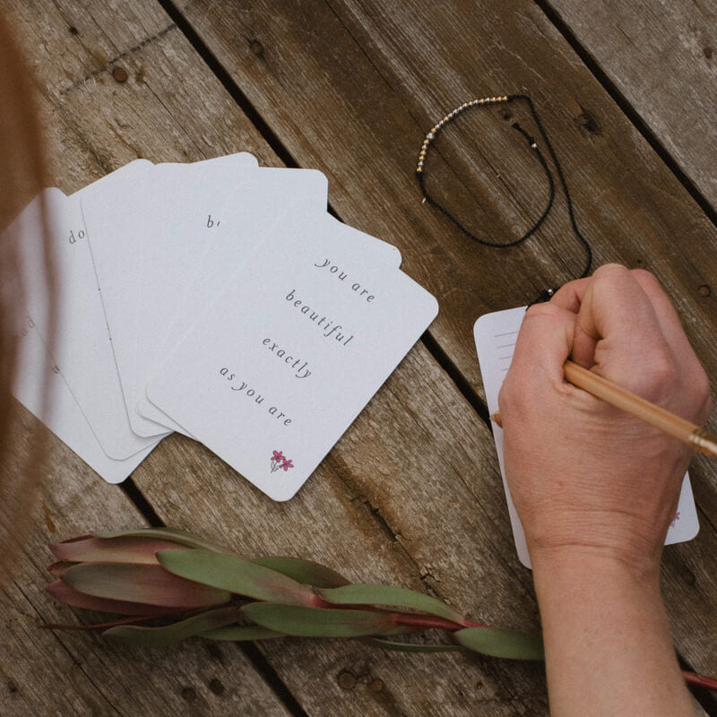 A woman writing on the back of Memory Makers Sweary Self-Love Edition. Some cards spread out next to her and a Strong AF Morse Code Bracelet