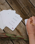 A woman writing on the back of Memory Makers Sweary Self-Love Edition. Some cards spread out next to her and a Strong AF Morse Code Bracelet