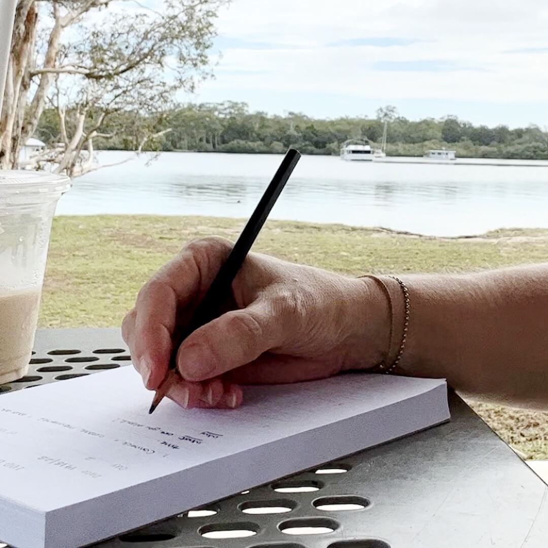 Woman&#39;s hand writing on 100 Days of 3 Things Notepad with a view of a river and an iced latte
