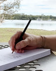 Woman's hand writing on 100 Days of 3 Things Notepad with a view of a river and an iced latte